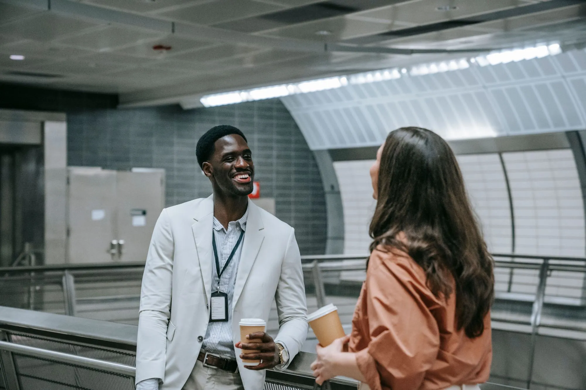 Zwei Menschen mit Kaffeebechern im Gespräch in einer U-Bahn-Station, einer lacht